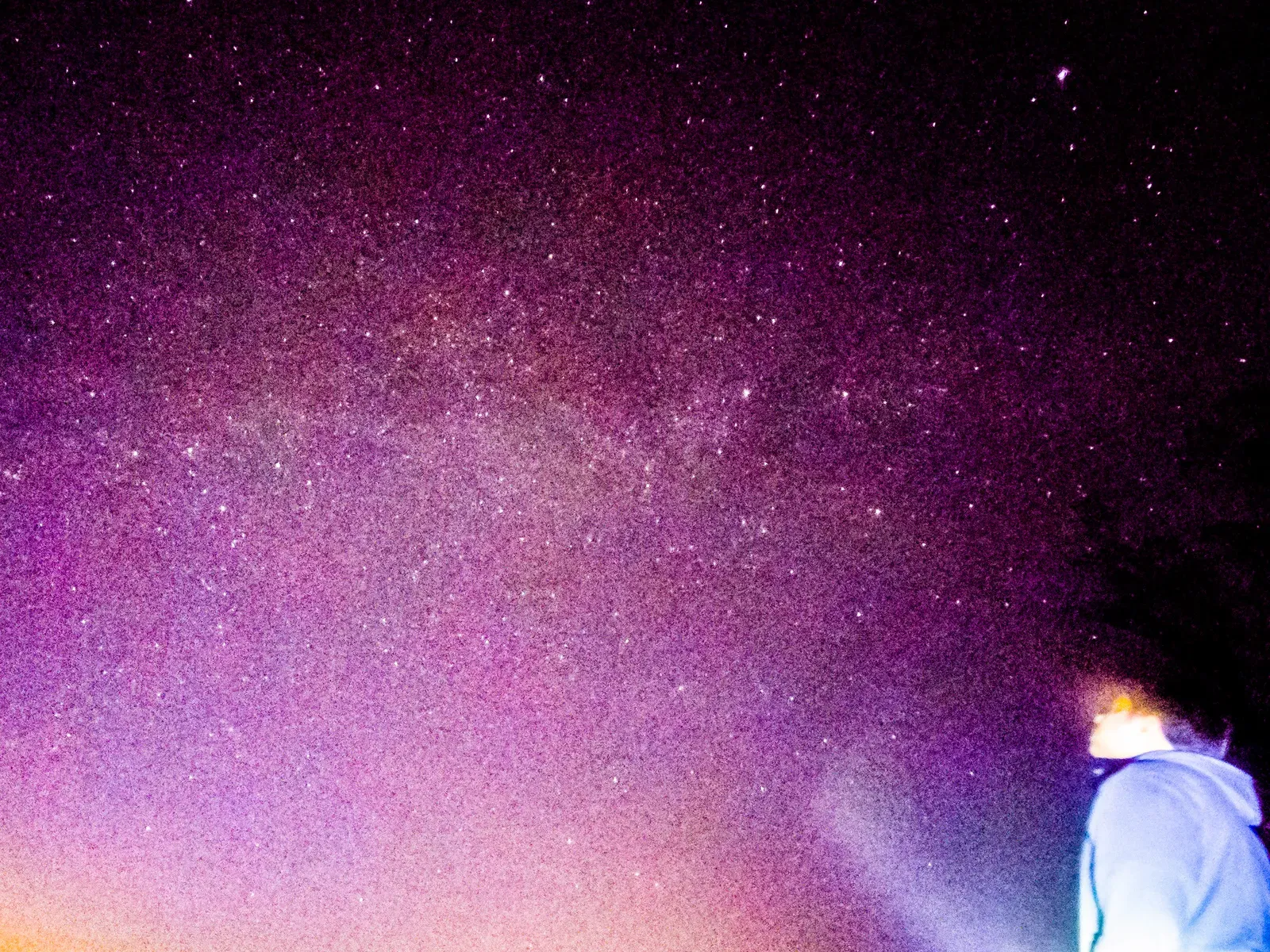 A grainy long-exposure of a star-filled night sky with a hooded figure in the foreground.